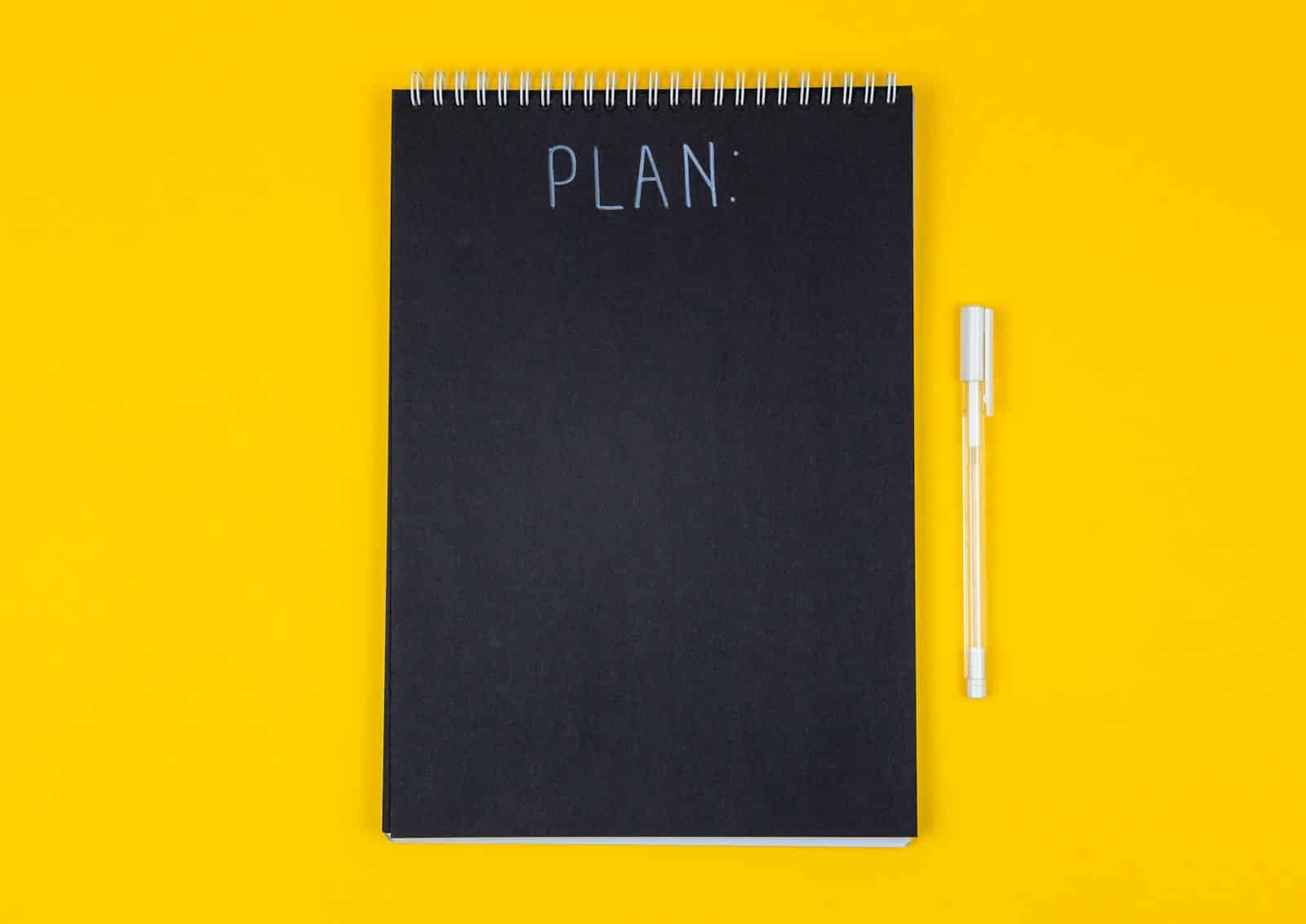 Carnet à spirale noir portant le mot PLAN sur fond jaune, pour préparer un cahier des charges de site web